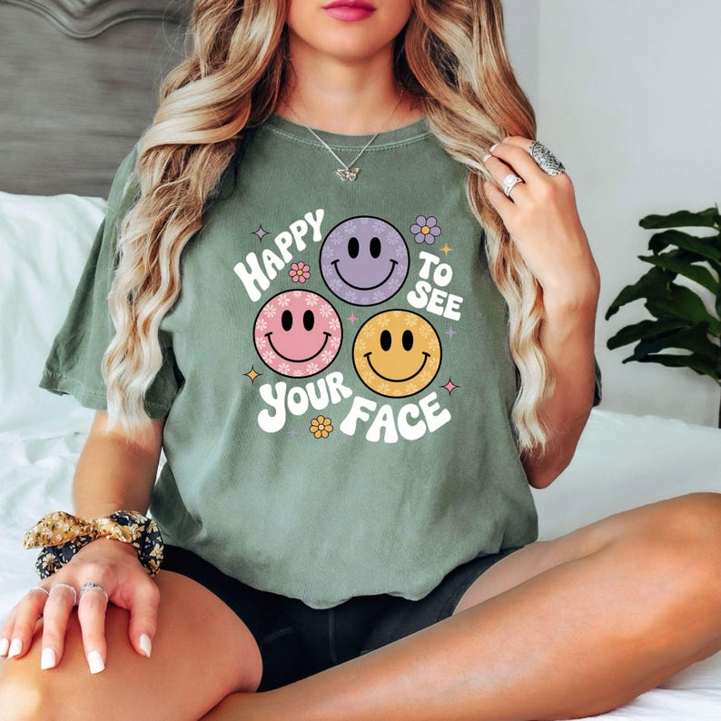 以下が含まれることがあります： 「Happy to See Your Face」の文字と、ピンク、紫、黄色のスマイリーフェイスのグラフィックがプリントされたセージグリーンのTシャツ。 ゆったりとしたフィット感で、半袖です。