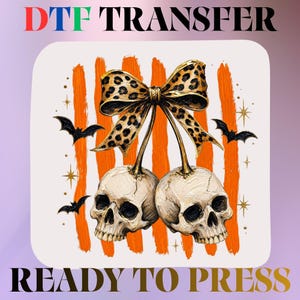 May include: A DTF transfer ready to press featuring two off-white skulls topped with a large leopard print bow. The design includes vertical orange brushstrokes, small black bats, and gold star accents. The text DTF TRANSFER and READY TO PRESS appears.