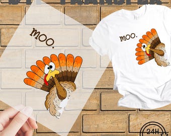 以下が含まれることがあります： 漫画の七面鳥のデザインと「moo」という言葉が入った白いTシャツ。七面鳥はオレンジと茶色の羽、黄色のくちばし、赤い肉垂れがあります。画像には、同じデザインのプレスする準備ができた転写も表示されています。
