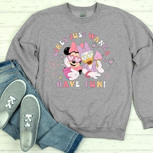 Op de afbeelding: Een grijze sweatshirt met een afbeelding van Minnie Mouse en Daisy Duck, met de tekst "GIRLS JUST WANNA HAVE FUN!". Minnie Mouse draagt een hartvormige zonnebril en houdt een roze beker vast. Daisy Duck houdt een roze ijsje vast.