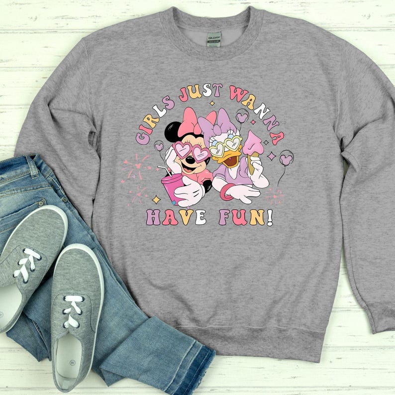 Op de afbeelding: Een grijze sweatshirt met een afbeelding van Minnie Mouse en Daisy Duck, met de tekst "GIRLS JUST WANNA HAVE FUN!". Minnie Mouse draagt een hartvormige zonnebril en houdt een roze beker vast. Daisy Duck houdt een roze ijsje vast.