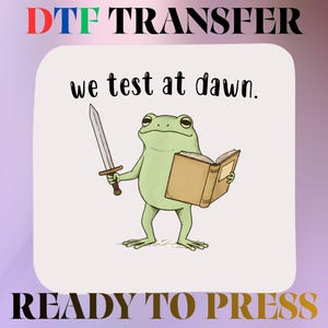 May include: A DTF transfer ready to press featuring a light green frog holding a silver sword in one hand and an open brown book in the other. Above the frog, the text reads we test at dawn. The design is set against a white background.