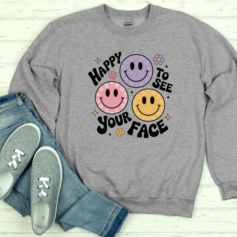 以下が含まれることがあります： 「Happy to See Your Face」の文字と、ピンク、紫、黄色のスマイリーフェイスが3つプリントされたグレーのスウェットシャツ。青いジーンズとグレーのスニーカーと合わせています。