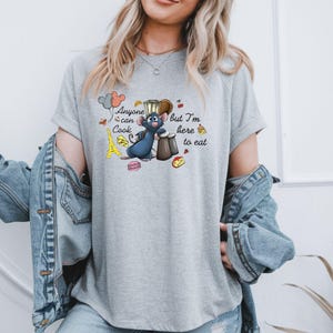 May include: A light gray t-shirt featuring a cartoon illustration of a blue rat chef with the text "Anyone can Cook but I'm here to eat." The design includes a miniature Eiffel Tower, macarons, and cheese.