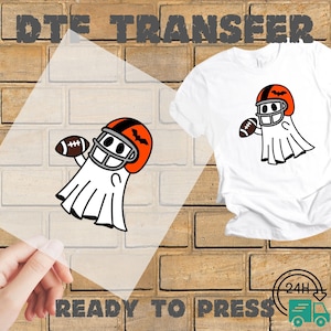 May include: A white t-shirt with a graphic of a ghost wearing a football helmet and holding a football. The helmet is orange and black. The words "DTF TRANSFER" and "READY TO PRESS" are printed on the image.