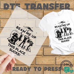 May include: A white t-shirt and a clear transfer sheet with a black graphic design. The design features a retro image of people being beamed up by a UFO, with the text "BEAM ME UP, THIS PLACE SUCKS!" The words "DTS TRANSFER" and "READY TO PRESS" are also visible.
