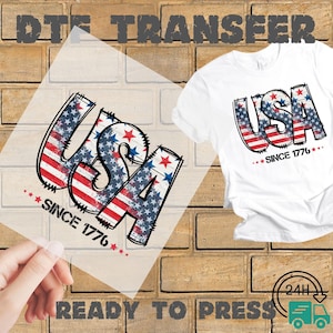 May include: A white t-shirt with a patriotic "USA" design, featuring red and blue stars and stripes, and the text "SINCE 1776". A clear transfer sheet with the same design is held up next to the shirt. The words "DTF TRANSFER" and "READY TO PRESS" are also visible.