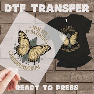 May include: DTF transfer featuring a butterfly design with the text "Nolite Te Bastardes Carborundorum." The design is in gold and black, ready to press onto a black t-shirt. The words "Ready to Press" are also visible.