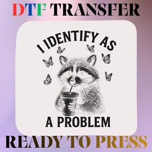May include: A DTF transfer sheet featuring a black and white sketch of a raccoon holding a drink cup, surrounded by butterflies. The text reads I IDENTIFY AS A PROBLEM. The design is on a light background with the words DTF TRANSFER READY TO PRESS.