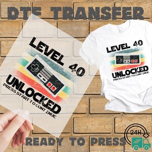 May include: A white t-shirt with a retro video game controller design. The design includes the text "LEVEL 40 UNLOCKED" with a colorful striped background. A clear transfer sheet with the same design is held in front of the shirt.