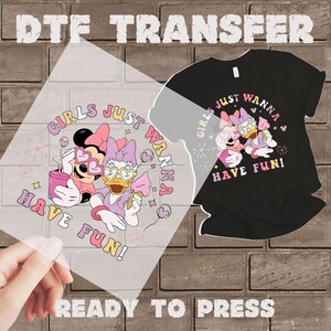 Op de afbeelding: Een DTF-transfer met Minnie Mouse en Daisy Duck met de tekst "GIRLS JUST WANNA HAVE FUN!" op een transparant vel. Er wordt ook een zwart t-shirt met hetzelfde ontwerp getoond. De woorden "READY TO PRESS" staan onderaan.
