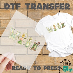 May include: White t-shirt with a colorful fairy design, alongside a clear DTF transfer sheet featuring the same design. The design includes fairies, bottles, and a mushroom house. The words "DTF TRANSFER" and "READY TO PRESS" are visible.