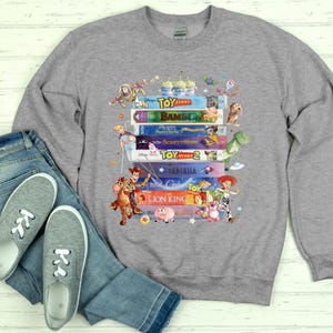 May include: A gray sweatshirt featuring a stack of classic Disney VHS tapes, including "Toy Story," "Bambi," and "The Lion King." Colorful cartoon characters like Woody, Buzz Lightyear, and others surround the tapes. The sweatshirt is paired with blue jeans and gray sneakers.