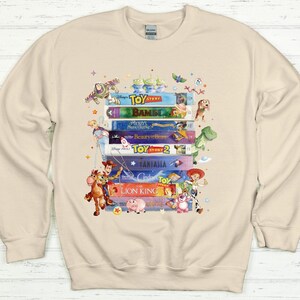 May include: Beige sweatshirt featuring a stack of colorful VHS tapes with Disney movie titles like "Toy Story," "Bambi," and "The Lion King." The design includes cartoon characters from the movies, such as Woody, Buzz Lightyear, and Simba.