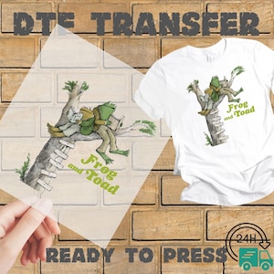 May include: White t-shirt with a green and brown illustration of Frog and Toad sitting on a tree branch, reading books. The words "Frog and Toad" are printed in green below the image. A DTE transfer is shown next to the shirt.