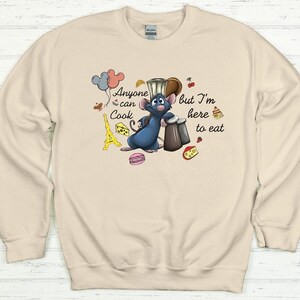 May include: Beige sweatshirt featuring a cartoon rat chef with the text "Anyone can Cook but I'm here to eat." The design includes balloons, a cheese wedge, a macaron, and the Eiffel Tower.