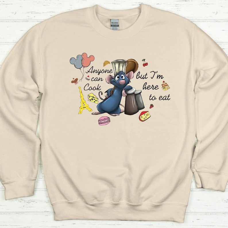 May include: Beige sweatshirt featuring a cartoon rat chef with the text "Anyone can Cook but I'm here to eat." The design includes balloons, a cheese wedge, a macaron, and the Eiffel Tower.