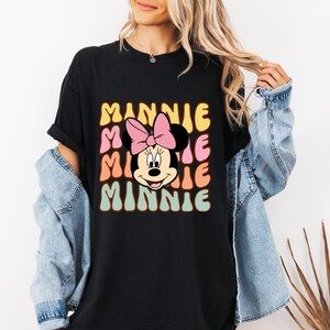 Może przedstawiać: Czarny t-shirt z retro wzorem Myszki Minnie i napisem "MINNIE". Projekt zawiera r&oacute;żową kokardę i napis "MINNIE" w kolorze ż&oacute;łtym, r&oacute;żowym, pomarańczowym i zielonym.