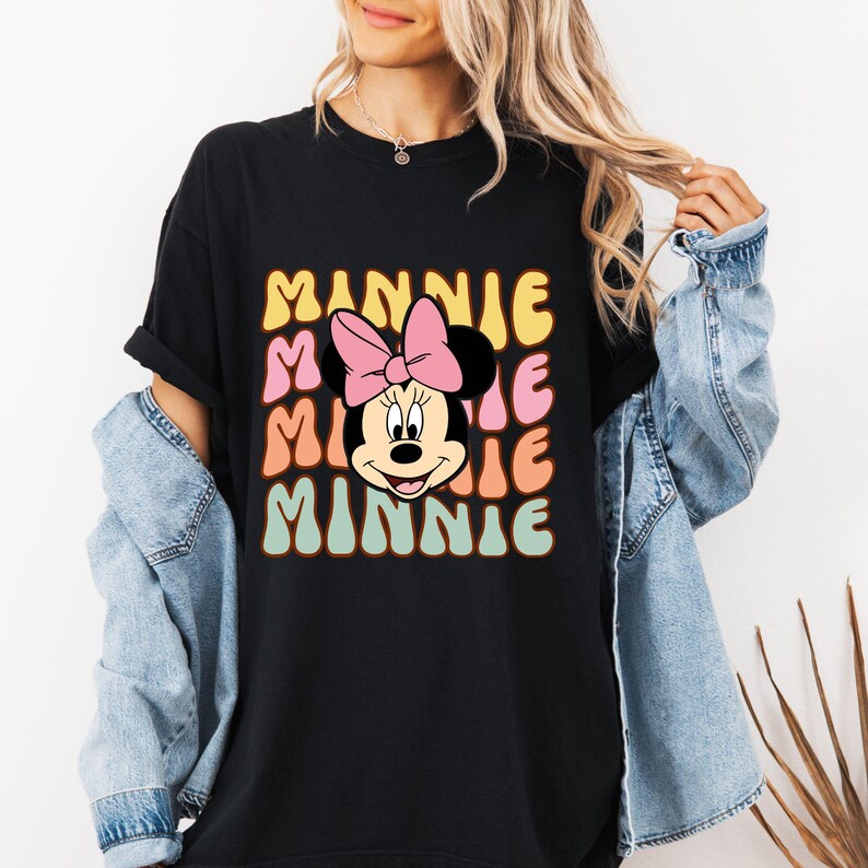 Może przedstawiać: Czarny t-shirt z retro wzorem Myszki Minnie i napisem "MINNIE". Projekt zawiera r&oacute;żową kokardę i napis "MINNIE" w kolorze ż&oacute;łtym, r&oacute;żowym, pomarańczowym i zielonym.