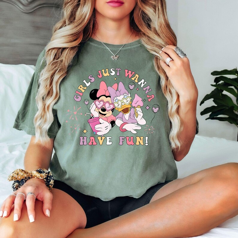 Op de afbeelding: Olijfgroen T-shirt met de tekst "GIRLS JUST WANNA HAVE FUN!" met Minnie Mouse en Daisy Duck. Minnie draagt een hartvormige zonnebril en Daisy draagt een zonnebril. Beide personages houden lekkernijen vast.