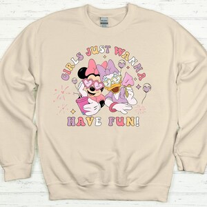 Op de afbeelding: Beige sweatshirt met een afbeelding van Minnie Mouse en Daisy Duck met een hartvormige zonnebril. De tekst "GIRLS JUST WANNA HAVE FUN!" is gedrukt in een kleurrijk, retro lettertype. De sweatshirt heeft een ronde hals en lange mouwen.