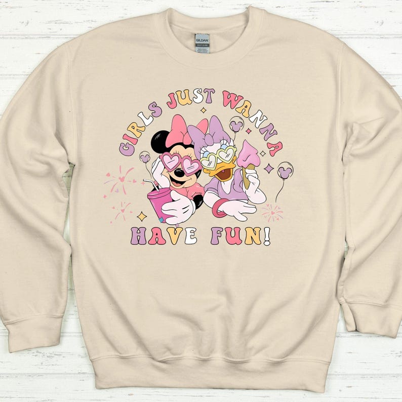 Op de afbeelding: Beige sweatshirt met een afbeelding van Minnie Mouse en Daisy Duck met een hartvormige zonnebril. De tekst "GIRLS JUST WANNA HAVE FUN!" is gedrukt in een kleurrijk, retro lettertype. De sweatshirt heeft een ronde hals en lange mouwen.