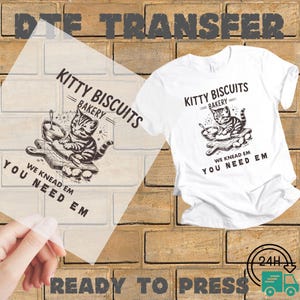 May include: White t-shirt with a black and white graphic of a cat kneading dough, with the text "Kitty Biscuits Bakery". The phrase "We Knead Em, You Need Em" is also printed on the shirt. A DTF transfer is shown.