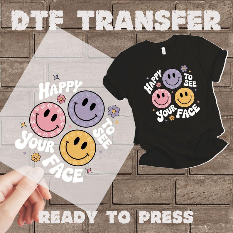以下が含まれることがあります： 「Happy to See Your Face」の文字が入った黒いTシャツと、同じデザインのDTF転写シート。デザインは、ピンク、紫、黄色の3つのスマイリーフェイスと花のアクセントが特徴です。「DTF TRANSFER」と「READY TO PRESS」の文字も表示されています。