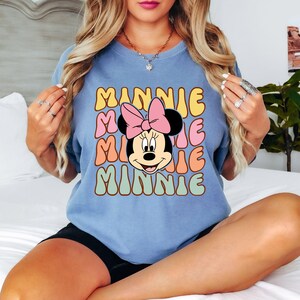 Może przedstawiać: Jasnoniebieska koszulka z retro wzorem. Projekt przedstawia imię "MINNIE" w ułożonej, kolorowej czcionce oraz kresk&oacute;wkowy wizerunek Myszki Minnie z r&oacute;żową kokardą.