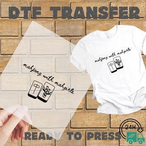 May include: A white t-shirt and a clear DTF transfer sheet, both featuring a black graphic of two mahjong tiles and the text "mahjong with mahgirls" in a cursive font. The tiles display a single character and a bamboo stalk design.