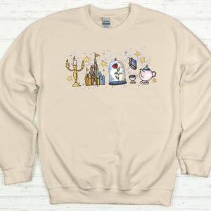 K&ouml;nnte beinhalten: Cremefarbenes Sweatshirt mit einer skurrilen Illustration mit Schloss, Rose in einer Glaskuppel, Teekanne und anderen m&auml;rchenhaften Elementen. Das Design enth&auml;lt goldene, blaue und gelbe Akzente und erzeugt eine charmante und verspielte &Auml;sthetik.