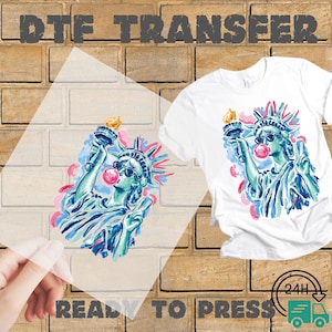 May include: DTF transfer featuring a colorful illustration of the Statue of Liberty wearing sunglasses and blowing a bubblegum bubble. The design is on a white t-shirt and a clear transfer sheet. Text reads "DTF TRANSFER" and "READY TO PRESS".