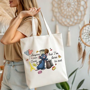 May include: Cream-colored tote bag featuring a cartoon illustration of a blue rat chef with the text "Anyone can cook but I'm here to eat." The design includes a chef's hat, Eiffel Tower, cheese, and macarons.