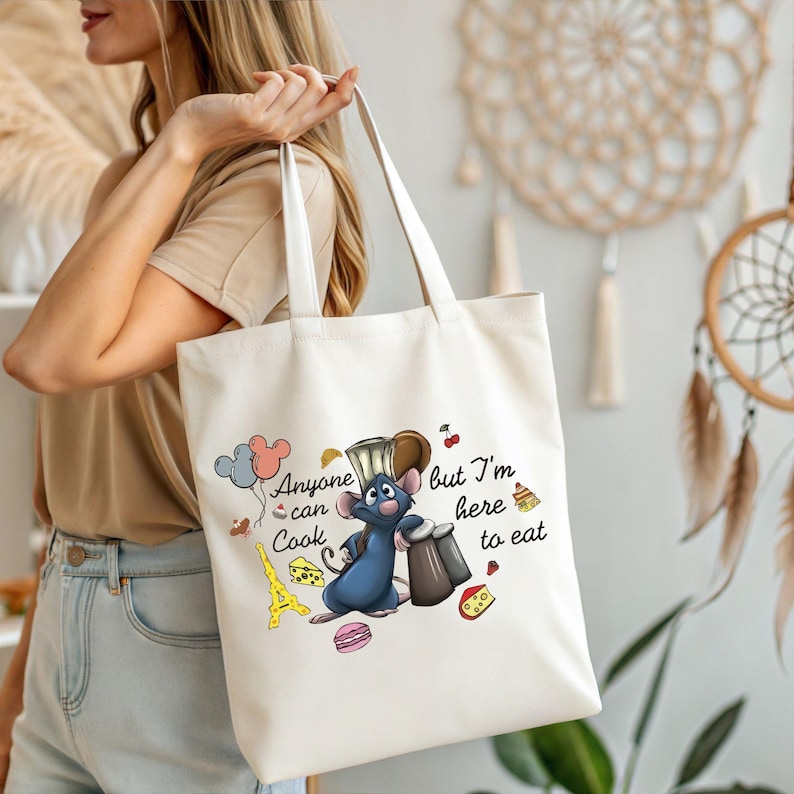 May include: Cream-colored tote bag featuring a cartoon illustration of a blue rat chef with the text "Anyone can cook but I'm here to eat." The design includes a chef's hat, Eiffel Tower, cheese, and macarons.