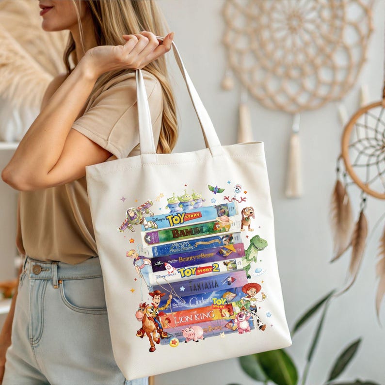 May include: Cream-colored tote bag featuring a stack of colorful VHS tapes with popular animated movie titles and characters. The bag has white handles and is held by a person. The design includes characters from Toy Story, Bambi, and The Lion King.