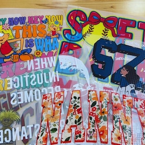 May include: A collection of heat transfer vinyl sheets featuring various designs. Included are a deer in a winter scene, cartoon characters with text, floral letters spelling "MAMA", and a baseball-themed design with the word "SOFT".