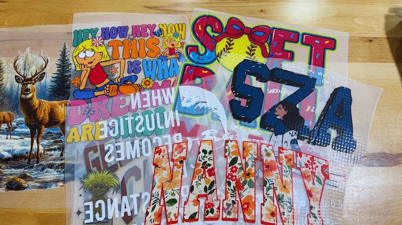 May include: A collection of heat transfer vinyl sheets featuring various designs. Included are a deer in a winter scene, cartoon characters with text, floral letters spelling "MAMA", and a baseball-themed design with the word "SOFT".