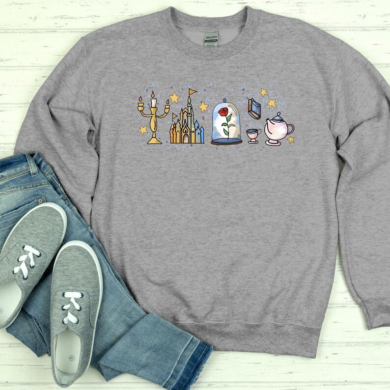 K&ouml;nnte beinhalten: Graues Sweatshirt mit einem farbenfrohen Cartoon-Motiv, das einen Kerzenleuchter, ein Schloss, eine Rose in einer Glaskuppel, ein Buch, eine Teekanne und eine Tasse zeigt. Das Design hat gelbe Sterne.