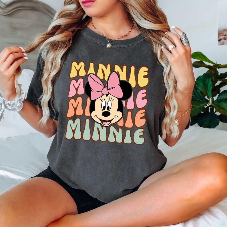 Może przedstawiać: Ciemnoszara koszulka z napisem "MINNIE" ułożonym czterokrotnie w stylu retro i grafiką Myszki Minnie. Projekt wykorzystuje kolory ż&oacute;łty, r&oacute;żowy, pomarańczowy i miętowy. To element odzieży codziennej.