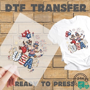 May include: A DTF transfer sheet and a white t-shirt featuring a graphic of two chipmunks wearing patriotic clothing, holding balloons and a flag, with fireworks and the text USA. The design uses red, white, and blue colors throughout the illustration.