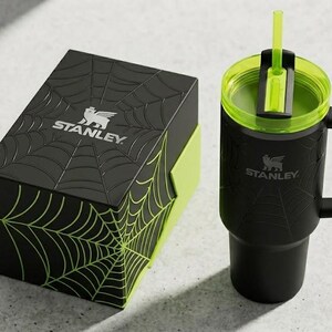 May include: Black Stanley tumbler with a green lid and straw, next to a matching black box. Both items feature a spiderweb design and the Stanley logo. The tumbler has a handle.