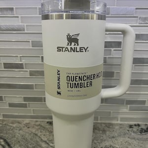 K&ouml;nnte beinhalten: Ein wei&szlig;er Stanley Quencher H2O Becher mit Griff. Der Becher hat einen transparenten Deckel und ein hellgr&uuml;nes Etikett mit dem Text "THE FLOWSTATE QUENCHER H2O TUMBLER" und "40 OZ | 1,18 L". Das Stanley-Logo ist sichtbar.