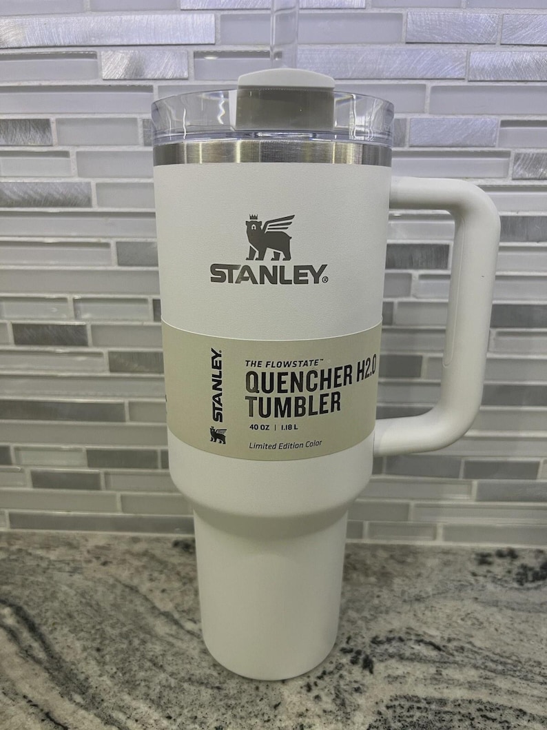 K&ouml;nnte beinhalten: Ein wei&szlig;er Stanley Quencher H2O Becher mit Griff. Der Becher hat einen transparenten Deckel und ein hellgr&uuml;nes Etikett mit dem Text "THE FLOWSTATE QUENCHER H2O TUMBLER" und "40 OZ | 1,18 L". Das Stanley-Logo ist sichtbar.