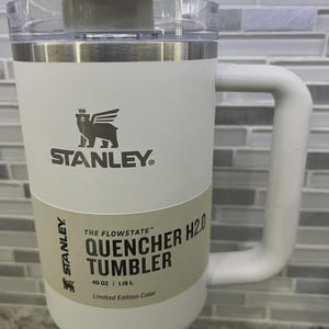 K&ouml;nnte beinhalten: Ein wei&szlig;er Stanley Quencher H2.0 Becher mit Griff und transparentem Deckel. Der Becher hat das Stanley-Logo und den Text "QUENCHER H2.0 TUMBLER" und "40 OZ | 1,18 L". Der Becher ist eine limitierte Edition.