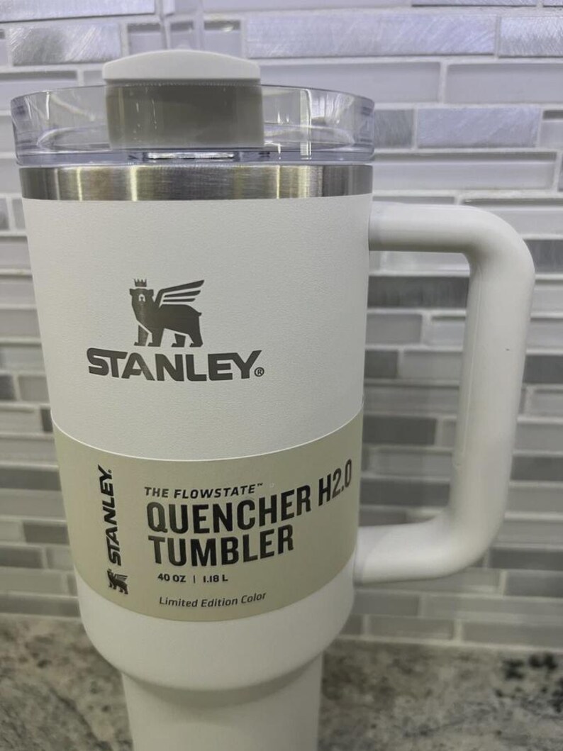 K&ouml;nnte beinhalten: Ein wei&szlig;er Stanley Quencher H2.0 Becher mit Griff und transparentem Deckel. Der Becher hat das Stanley-Logo und den Text "QUENCHER H2.0 TUMBLER" und "40 OZ | 1,18 L". Der Becher ist eine limitierte Edition.