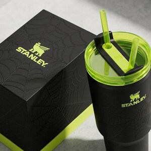 May include: Black Stanley tumbler with a green lid and straw, and a matching box. The tumbler and box feature the Stanley logo in green and a spiderweb design. The tumbler has a handle.