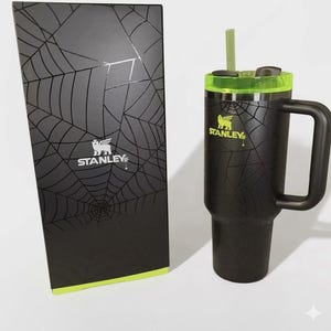May include: Black Stanley tumbler with a handle, green accents, and a straw. The tumbler features a spiderweb design and the Stanley logo. A matching black box with a spiderweb design and the Stanley logo is next to the tumbler.