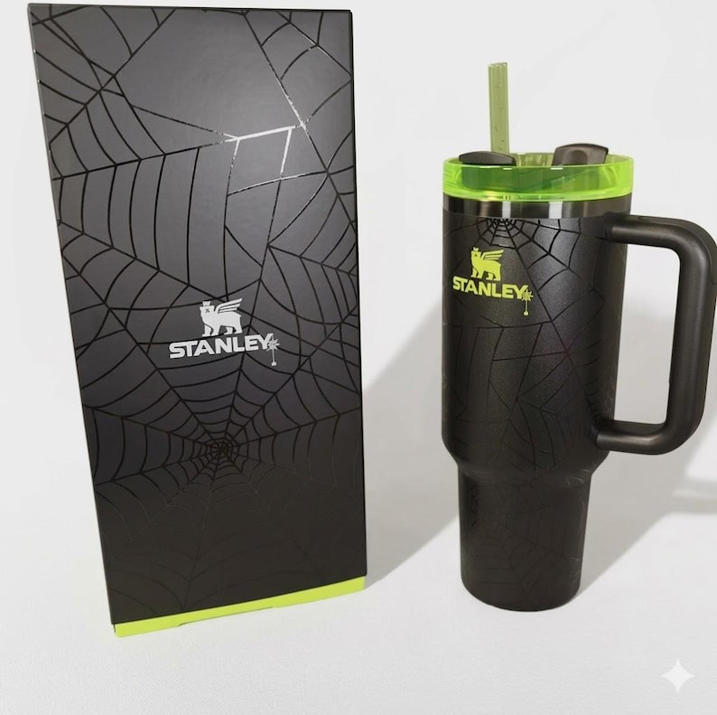May include: Black Stanley tumbler with a handle, green accents, and a straw. The tumbler features a spiderweb design and the Stanley logo. A matching black box with a spiderweb design and the Stanley logo is next to the tumbler.