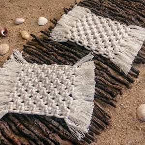 Könnte beinhalten: Zwei cremefarbene Makramee-Untersetzer mit Fransen an den Kanten liegen auf einem Bett aus Zweigen, vor einem sandigen Hintergrund. Muscheln sind um die Untersetzer herum verstreut und erzeugen ein Strand-Thema. Die Untersetzer sind quadratisch und messen etwa 13 cm x 13 cm.
