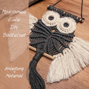 Könnte beinhalten: Handgefertigte Makramee-Eule zum Aufhängen in Grau und Creme. Die Eule hat große kreisförmige Augen und einen strukturierten Körper mit Holzdübeln und Fransen. Der Text "Makramee Eule DIY Bastelset" ist sichtbar.
